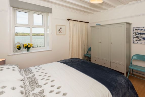 A bright bedroom featuring a bed, vintage wardrobe, and a view of water through a window.