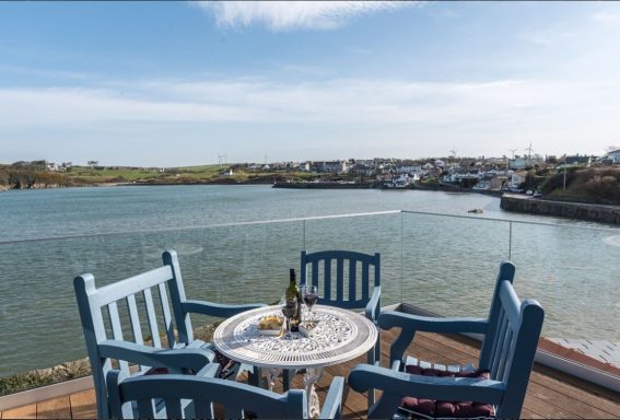 A coastal scene with a table, chairs, and a bottle overlooking a serene bay.