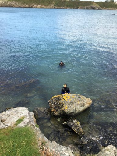 Two people in a calm body of water, with one sitting on a rock and the other swimming.