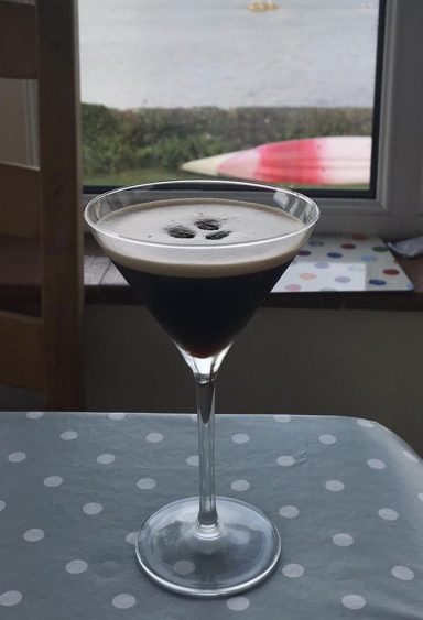 A martini glass filled with dark coffee topped with a creamy froth, set by a window.