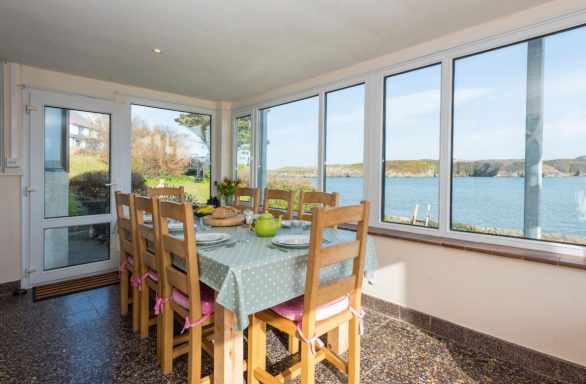 Bright dining area with a table, overlooking a scenic waterfront view through large windows.