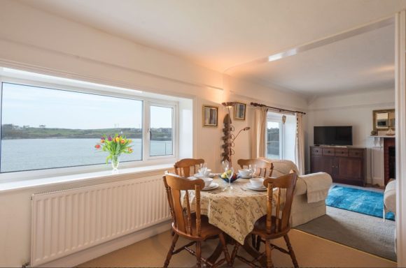 Bright, cozy room with dining table, flowers, and view of water through large window.