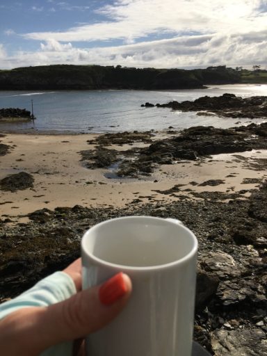A hand holding a white mug in front of a coastal scene with rocks and water.